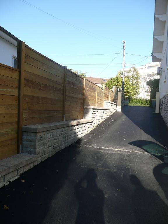 Pare-vue plein dominant une allée bitumée depuis un mur en pierre, fabrication française SNMB — bois autoclave classe 4